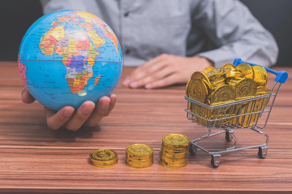 A person holds a globe beside stacked gold coins and a shopping cart filled with gold, symbolizing the commodities boom in 2025 with global investments in gold, silver, and oil.