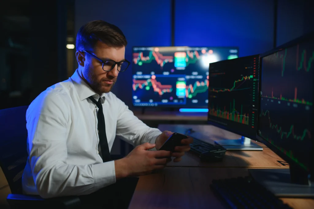 A forex trader analyzing live market charts on multiple screens while checking updates on his phone in a modern trading office.