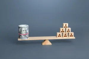 A roll of US dollar bills and wooden blocks with human icons balanced on a wooden seesaw, symbolizing economic power balance and De-Dollarization.