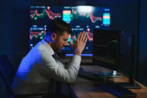 Frustrated trader sitting in front of multiple trading screens showing market charts and losses, illustrating FOMO in trading and emotional decision making.