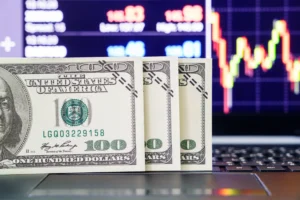 Three US hundred-dollar bills placed on a laptop keyboard with a bullish forex chart displayed in the background.