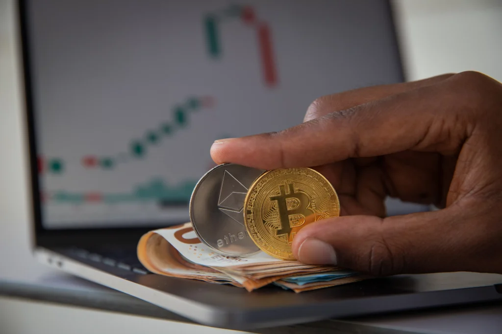 Hand holding Bitcoin and Ethereum coins with cash placed on a laptop showing crypto market charts.