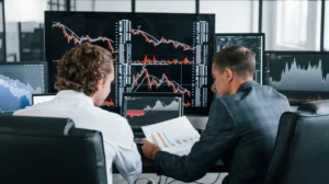 Two forex traders analyzing multiple currency charts on large trading screens to study multi pair correlation trading patterns.