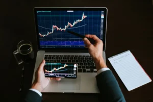 A trader analyzing cryptocurrency price charts on a laptop and smartphone, with a notepad showing trading notes and a cup of coffee nearby.