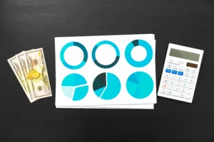Flat lay of financial analysis setup showing dollar bills, coins, printed pie charts in blue, and a calculator on a dark desk.