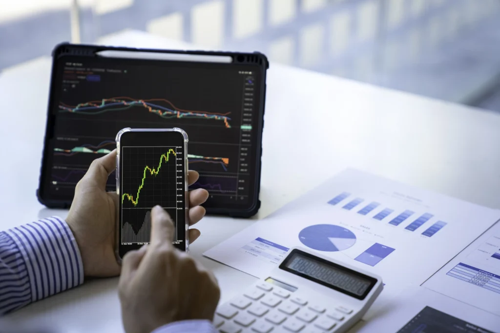 A person analyzing rising market charts on a smartphone and tablet with financial reports and a calculator on the desk.