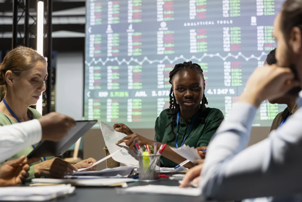 Team of financial analysts reviewing charts and market data during after-hours trading session with stock boards in background.